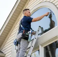 Professional window cleaner on a ladder using a squeegee to clean an exterior arched window, Miami, FL.